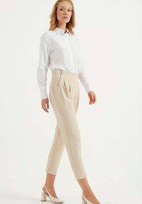 Beige plissé broek met een hoge taille, een zijrits en contrasterende stiksels, gecombineerd met een klassiek wit overhemd met knopen en hakken.