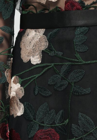 Zwarte doorschijnende stof met ingewikkelde bloemmotieven in beige, rood en groen, met een zwarte riem die de taille accentueert.