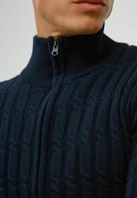 Mørkeblå strikket sweater med høj krave og lynlåset hals. Har en ribbet tekstur og et kabelstrikket mønster hele vejen igennem.