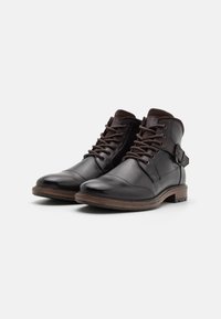 Botas de cuero marrón hasta el tobillo con un acabado suave, punta redondeada y diseño de cordones. Presenta una correa decorativa en el costado y una suela de madera.