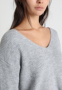 Grå stickad tröja med en bred V-ringning, med texturerad ribbning och en avslappnad passform. Tyget verkar mjukt och lätt.