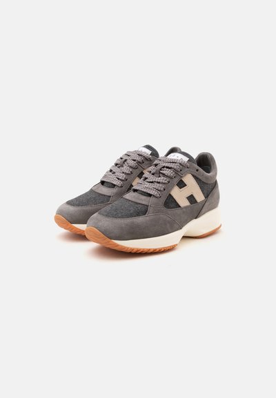 Paire de baskets grises en suède et tissu avec lacets gris, logo latéral beige, semelles intermédiaires blanches et semelles extérieures texturées marron clair sur fond blanc.
