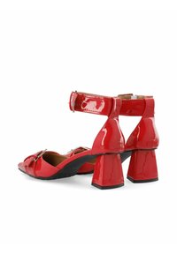 Sandalias de tacón de charol rojo con tira en el tobillo, tacones cuadrados y detalle decorativo con hebilla en la punta. Textura suave y brillante.