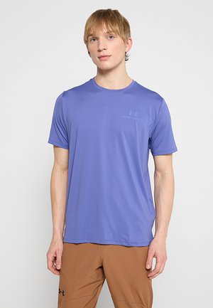 Młody mężczyzna w fioletowej koszulce z krótkim rękawem Under Armour i brązowych spodniach, stojący na jednolitym białym tle.
