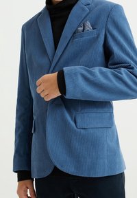 Modrý kordový blazer s vertikálním ribbovaným vzorem, jednou náprsní kapsou s vzorovaným kapesníčkem a klopami. Černý rolák pod ním.