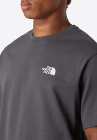 Mand iført en mørkegrå The North Face t-shirt, der viser logoet på venstre bryst, med fokus på overkrop og nakke.