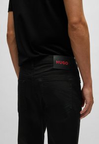 Sorte denim jeans med synlige sømme, baglommer og en rektangulær sort label med "HUGO" i fed rød tekst.