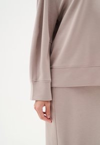 Personne portant un haut taupe à manches longues avec larges poignets et une jupe assortie, main détendue le long du corps sur un fond uni.