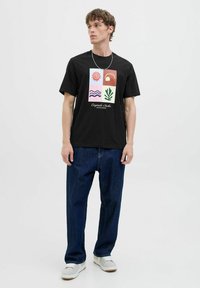 Schwarzes T-Shirt mit farbenfrohem Grafikdesign, das Sonne, Wellen, Pflanze und Hintergrundformen zeigt. Kombiniert mit blauen Jeans und weißen Turnschuhen.