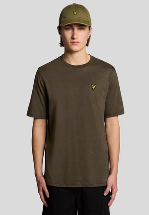 Jonge man met een olijfgroene pet en bijpassend T-shirt met een klein geel vogellogo op de borst, staande tegen een eenvoudige grijze achtergrond.