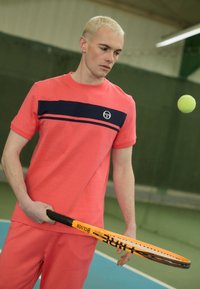 Korálové sportovní tričko s námořnickou vodorovnou pruhou a logem, spárováno s korálovými šortkami. Drží černou tenisovou raketu s oranžovým akcentem.