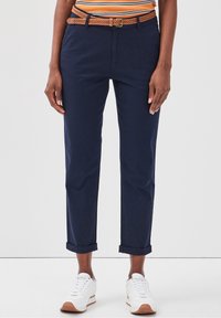 Pantalon en coton bleu marine avec un revers, associé à une ceinture tressée couleur tabac. Des baskets blanches avec des accents marron complètent la tenue.