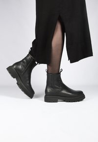 Botas de cuero negro hasta el tobillo con suela gruesa, cremallera lateral y tiradores. El diseño presenta una textura suave con un acabado mate.