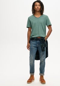 Grøn v-hals t-shirt, blå jeans, brune snøresko. Ternet skjorte bundet om livet, ur på håndleddet, tekstureret tøj, afslappet påklædning.