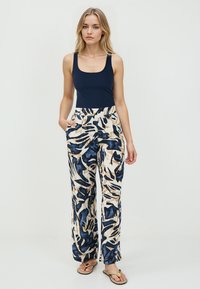 Jeune femme portant un haut sans manches bleu marine, un pantalon beige et bleu marine à motifs abstraits, et des sandales beiges, se tenant devant un fond uni.