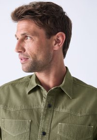 Green textured button-up shirt with two chest pockets and a classic collar, featuring black buttons and a close-fit design.