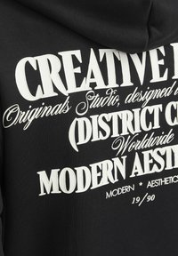Zwarte sweatshirt met grote, witte, gestileerde tekst die "CREATIVE," "DISTRICT," en "MODERN AESTHETIC" leest, met verschillende lettertypen en accenten.