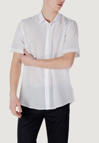 Camisa blanca de manga corta y botones, hecha de un tejido ligero, con color, tapeta delantera y puños enrollados. Textura suave, diseño minimalista.
