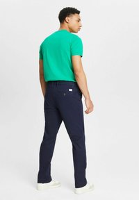 Zaļš kokvilnas T-krekls ar apaļu apkakli, apvienots ar tumši ziliem biksēm, kas ir ar aizmugurējām kabatām. Modelis stāv ar muguru vērsts, valkājot baltas sporta kurpes.