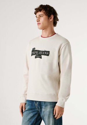 Svetlobéžová mikina s rebrovanými manžetami a lemom. Na prednej strane čierny grafický nápis "PEPE JEANS LONDON", nosená cez modré džínsy.