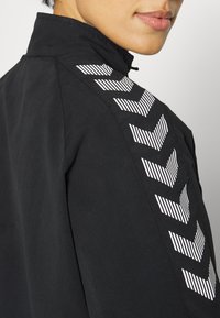 Svart jacka med hög krage, med ett geometriskt vitt randigt mönster på axeln och slät tygstruktur.