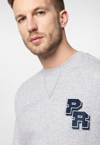 Szara bluza z okrągłym dekoltem, z naszytym granatowym logo "PR" na klatce piersiowej. Miękka tekstura i prosty design.