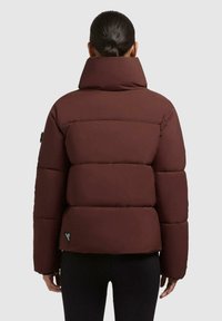 Donna con capelli scuri legati in una coda di cavallo, indossa un giubbotto invernale imbottito marrone con colletto alto, vista da dietro su uno sfondo chiaro e neutro.