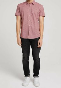 Camicia a maniche corte con bottoni, di colore rosa chiaro con un piccolo motivo geometrico, abbinata a jeans scuri e sneaker bianche.