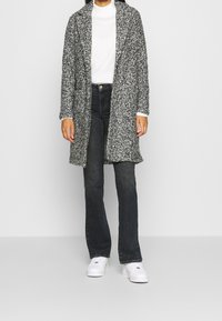 Grå och vit prickig lång cardigan, stickad textur, öppen fram, kombinerad med en vit polotröja och svarta utsvängda jeans, vita sneakers.