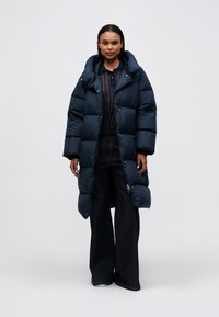 Žena stojící v dlouhém černém péřovém kabátu přes průhledný černý top a široké černé džíny na bílém pozadí.