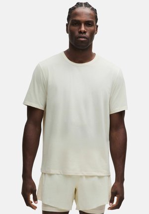 Mann mit geflochtenem Haar, der ein schlichtes hellbeiges Kurzarmhemd und passende Shorts trägt, steht vor weißem Hintergrund.