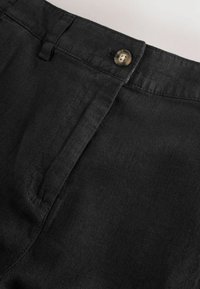 Schwarzer Hosenbund aus Stoff mit braunem Knopfverschluss und Gürtelschlaufe vor hellem Hintergrund.