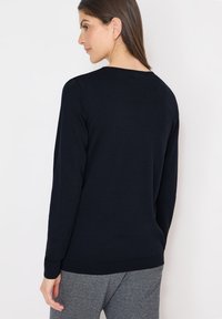 Femme aux cheveux bruns portant un pull noir uni à manches longues et un pantalon gris, de dos sur un fond blanc.