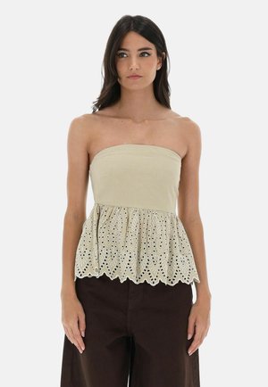 Giovane donna con capelli scuri che indossa un top beige senza spalline con ricamo a occhielli e pantaloni larghi scuri, in piedi davanti a uno sfondo semplice.