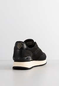 Czarny sneaker z teksturowanej tkaniny i skórzanym obcasem, beżową grubą podeszwą, widoczny z tyłu na szarym podłożu na jasnoszarym tle.