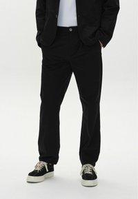 Black trousers made from smooth fabric, featuring a button closure and side pockets. Paired with black and white sneakers.