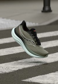 Αθλητικό παπούτσι σε ελιά πράσινο και μαύρο με παχιά λευκή σόλα να αιωρείται πάνω από μια διάβαση πεζών σε ασφαλτοστρωμένο δρόμο.