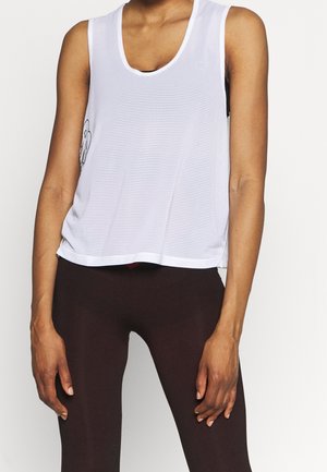 Kvinde iført en hvid net-top uden ærmer over en sort sports-bh og mørke leggings, stående med afslappede arme mod en ensfarvet baggrund.