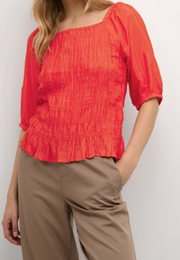 Blusa rossa, in tessuto strutturato, con scollatura quadrata, maniche a tre quarti e orlo arricciato, abbinata a pantaloni beige. Tessuto liscio con pieghe.