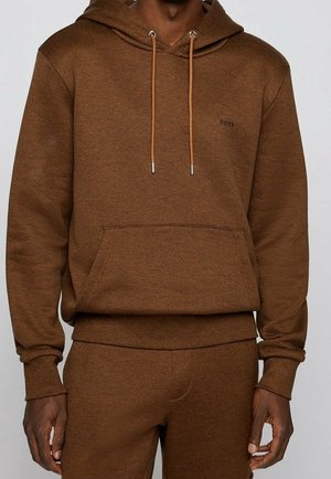 Mężczyzna ubrany w brązową teksturowaną bluzę z kapturem i pasujące spodnie dresowe z przednią kieszenią i sznurkami do ściągania.