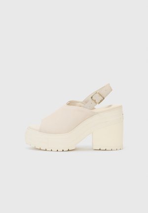 Platform heel sandal in beige suede with an open toe, adjustable ankle strap, and a chunky textured sole for stability.