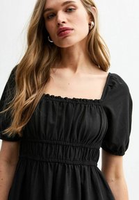 Vestido negro con escote cuadrado, mangas abullonadas y detalles fruncidos en la cintura. Textura de tela suave y silueta fluida.