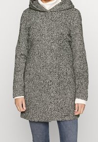 Abrigo de patrones en blanco y negro con capucha, presentado en una tela texturizada, de manga larga y con puños acanalados. Llevado sobre un cuello de tortuga blanco.