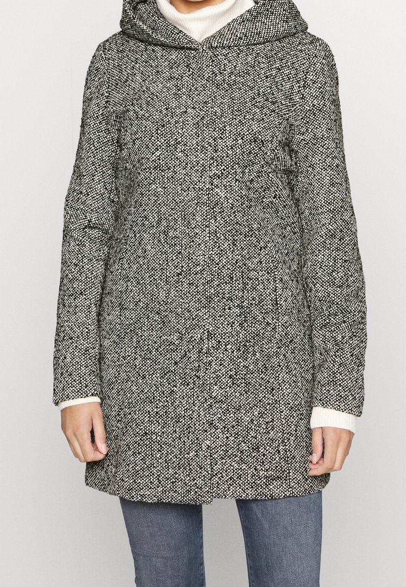 Abrigo de patrones en blanco y negro con capucha, presentado en una tela texturizada, de manga larga y con puños acanalados. Llevado sobre un cuello de tortuga blanco.