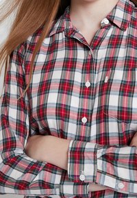 Camicia a quadri rossa, bianca e verde con bottoni; presenta un colletto, maniche lunghe e una piccola tasca sul petto con chiusura a bottone bianco.