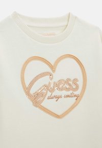 Felpa bianca con un design a forma di cuore. Paillette dorate formano la scritta "Guess" all'interno del cuore con "sempre sorridente" ricamato sotto. Tessuto strutturato.