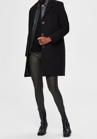 Schwarzer Wollmantel mit Revers, Knopfschluss und vorderen Taschen, getragen über einem grauen Hemd und olivgrünen Lederhosen mit schwarzen Stiefeletten.