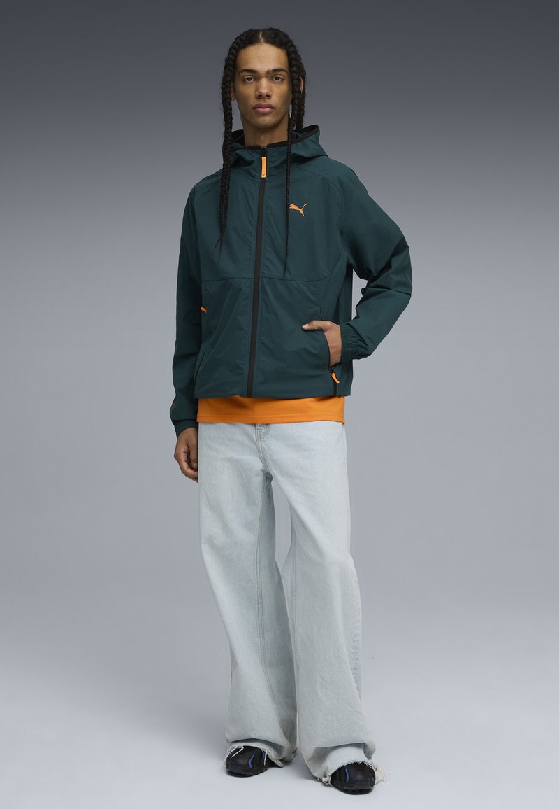 Chaqueta liviana de color verde oscuro con detalles en naranja, puños elásticos, dos bolsillos frontales, usada sobre una camiseta naranja y jeans de color azul claro.