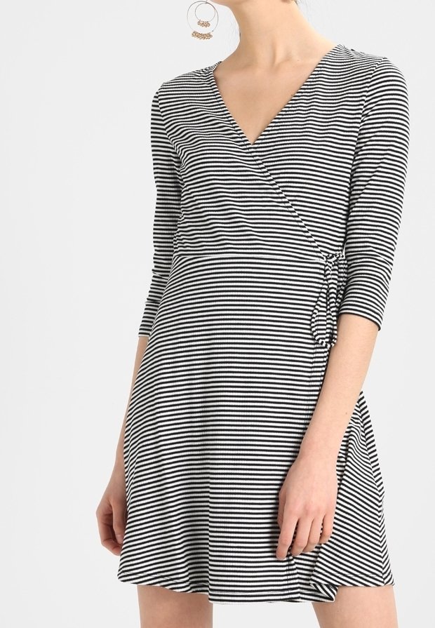 Femme portant une robe portefeuille rayée noire et blanche avec des manches trois-quarts et de grandes boucles d'oreilles, debout devant un fond clair.