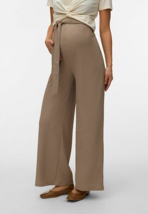 Donna che indossa pantaloni beige a gamba larga e vita alta con cintura in tessuto abbinata e scarpe piatte marroni, abbinati a una maglia color crema annodata.